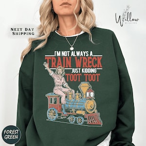 May include: Dark green sweatshirt featuring a graphic of a woman on a train, with the text "I'm not always a train wreck, just kidding, toot toot". The sweatshirt has a white collar.