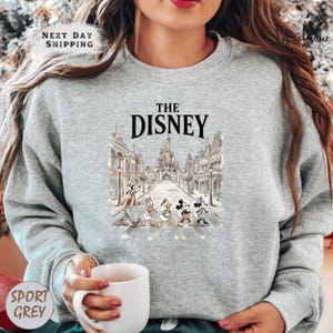 A woman wearing a gray sweatshirt with a graphic of Disney characters crossing an empty street.