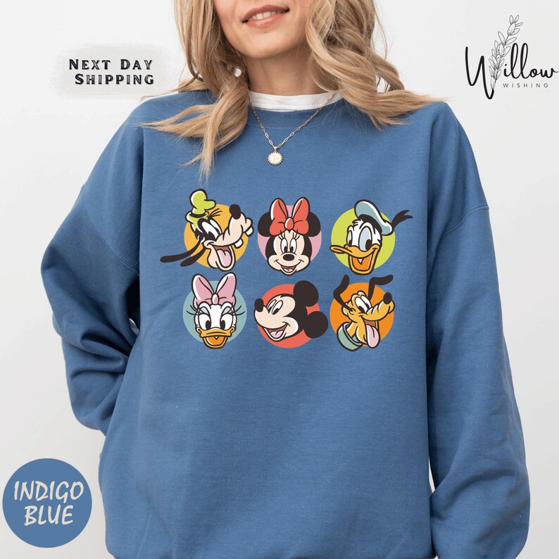 A woman wearing a blue sweatshirt with a graphic of six Disney character faces, including Minnie Mouse, Donald Duck, Mickey Mouse, Goofy, Donald Duck, and Pluto.