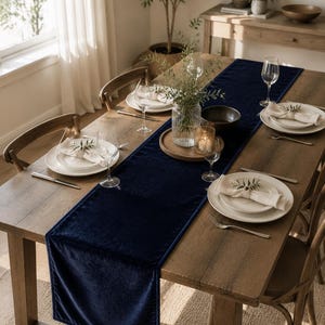 May include: A long, deep navy blue velvet table runner draped over a rustic wooden dining table. The runner features a smooth, soft-textured fabric with finished edges, centered beneath a glass vase, a wooden tray, and place settings.
