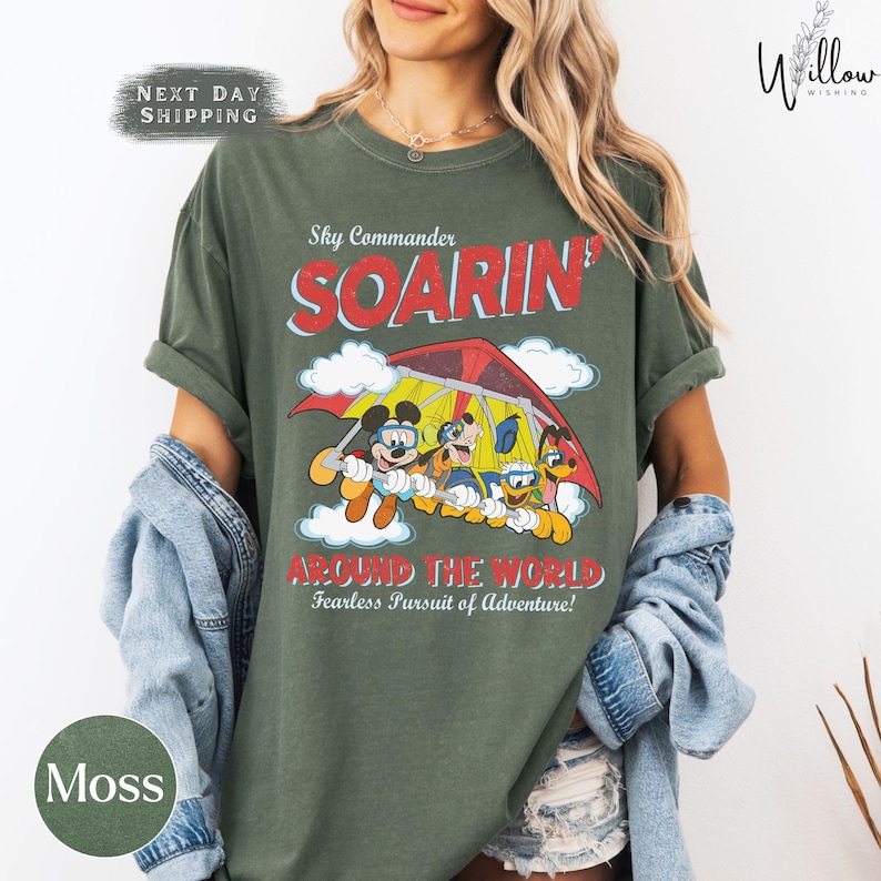 A woman wearing a green t-shirt with a cartoon image of Mickey Mouse and Goofy on a parachute, along with the text "Sky Commander SOARIN' AROUND THE WORLD Fearless Pursuit of Adventure!"