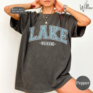 A person wearing a gray t-shirt with the word "LAKES" printed on it, along with the words "WEEKEND" below it. The person is posing with their hands on their head.