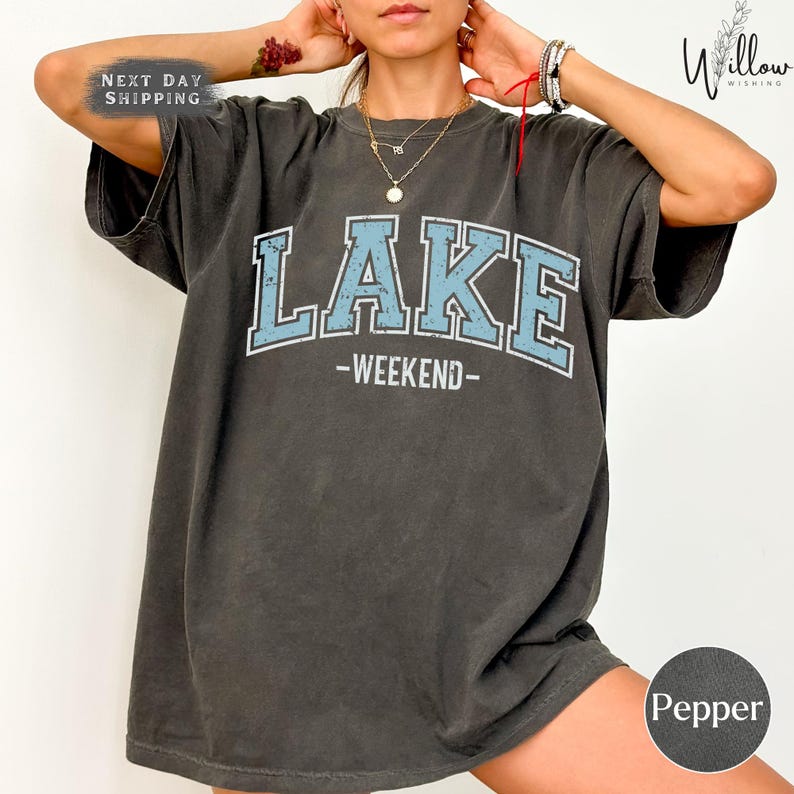 A person wearing a gray t-shirt with the word "LAKES" printed on it, along with the words "WEEKEND" below it. The person is posing with their hands on their head.