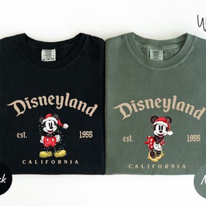 two t-shirts with the same design, featuring a Mickey Mouse character and text that reads "Disneyland est. 1955 California". The t-shirts are in different colors: black and green.