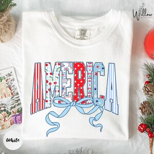 a white t-shirt with a patriotic design that says "AMERICA" in a stylized font, with a blue ribbon and red and white stripes. The t-shirt is laid out on a surface, surrounded by holiday decorations such as pine cones and ornaments.