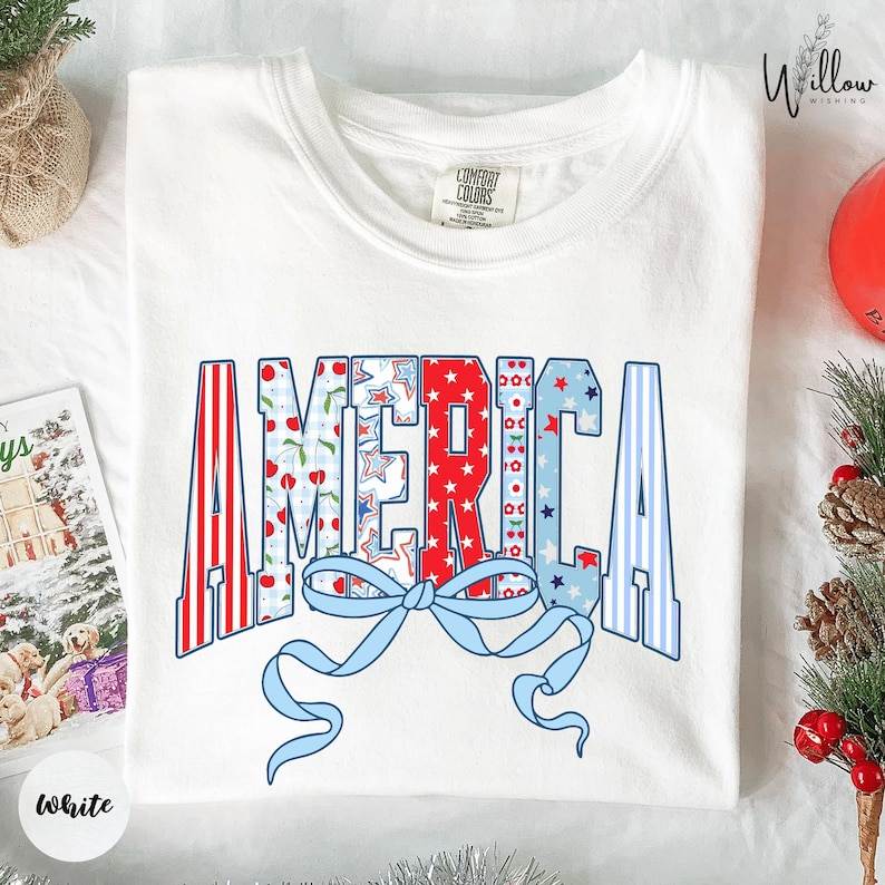 a white t-shirt with a patriotic design that says "AMERICA" in a stylized font, with a blue ribbon and red and white stripes. The t-shirt is laid out on a surface, surrounded by holiday decorations such as pine cones and ornaments.