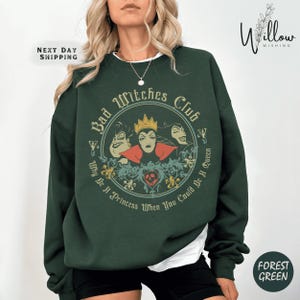 May include: Forest green sweatshirt with a circular graphic featuring three cartoon witches and the text "Bad Witches Club." The design includes the phrase "Why Be A Princess When You Could Be A Queen." The sweatshirt has a crew neck.