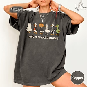 A person wearing a gray t-shirt with a Halloween-themed graphic of various cartoon animals, including a ghost, pumpkin, skeleton, and ducks, along with the text "Just a spooky goose".
