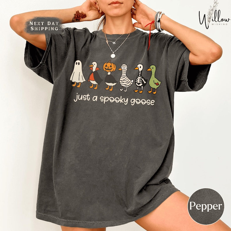 A person wearing a gray t-shirt with a Halloween-themed graphic of various cartoon animals, including a ghost, pumpkin, skeleton, and ducks, along with the text "Just a spooky goose".