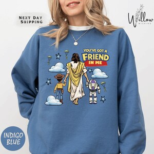 A woman wearing a blue sweatshirt with a cartoon illustration of a woman holding a child and a robot, along with the text "You've got a FRIEND in ME" and a Bible verse reference.