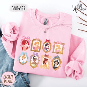 A pink sweatshirt with a design of various Disney princesses in frames, including Cinderella, Snow White, and Belle.