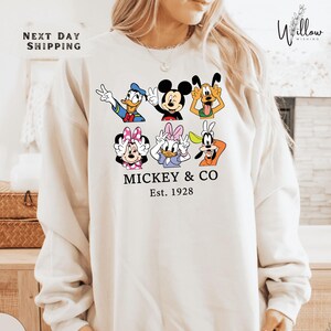 A woman wearing a white sweatshirt with a graphic design featuring various Disney characters such as Mickey Mouse, Donald Duck, Goofy, Minnie Mouse, and Pluto.