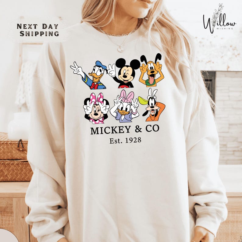 A woman wearing a white sweatshirt with a graphic design featuring various Disney characters such as Mickey Mouse, Donald Duck, Goofy, Minnie Mouse, and Pluto.
