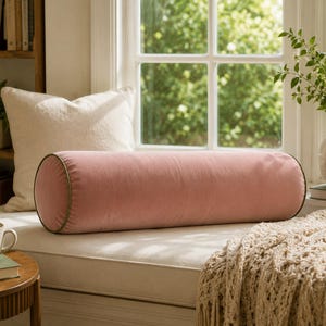 May include: A cylindrical bolster pillow in a soft dusty pink velvet fabric, featuring dark green piping along the circular edges. The pillow is elongated and maintains a firm, rounded shape, providing a solid color accent for home decor.
