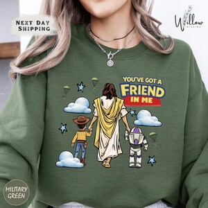 A green sweatshirt with a graphic design depicting a person in a yellow robe and a child, along with a character from the Toy Story franchise, and the text "You've got a FRIEND in ME".