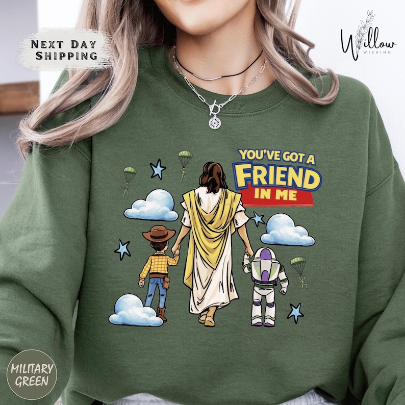 A green sweatshirt with a graphic design depicting a person in a yellow robe and a child, along with a character from the Toy Story franchise, and the text "You've got a FRIEND in ME".