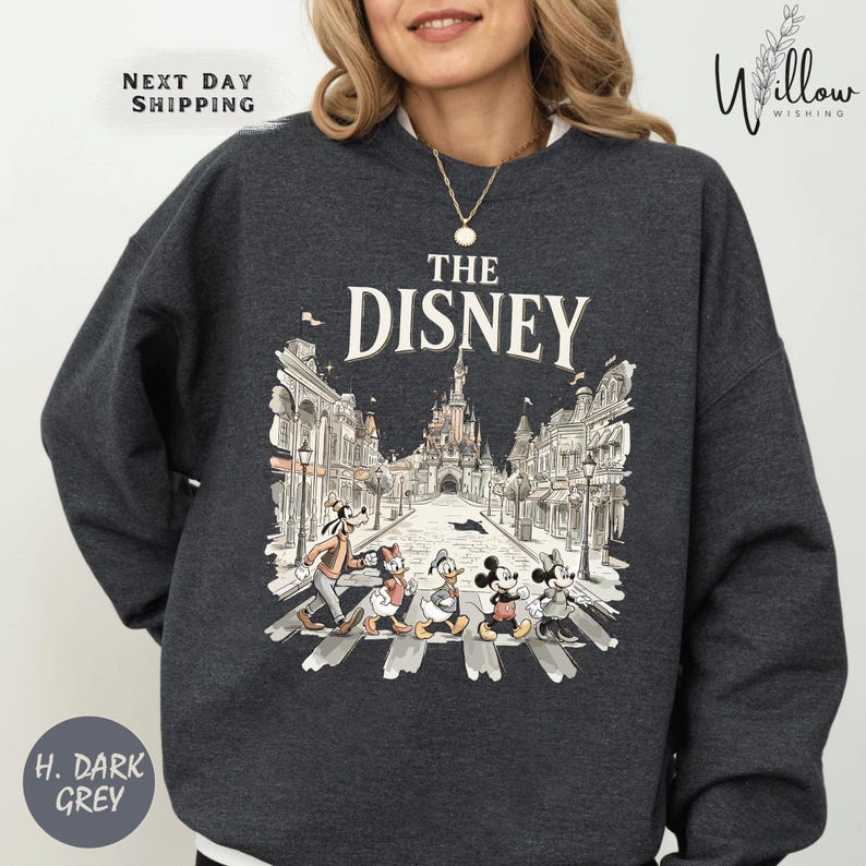 A woman wearing a gray sweatshirt with a graphic design featuring Disney characters crossing a street.
