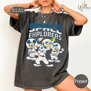 May include: Charcoal grey t-shirt with the text "SPACE EXPLORERS" in blue and white. The design shows Mickey Mouse, Goofy, and Donald Duck in astronaut suits. The t-shirt has a loose, comfortable fit.