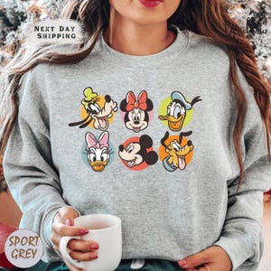 A woman wearing a gray sweatshirt with a graphic of Disney characters on it, holding a white coffee mug.