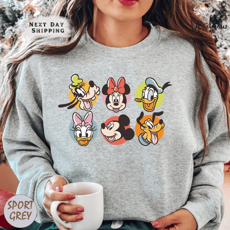 A woman wearing a gray sweatshirt with a graphic of Disney characters on it, holding a white coffee mug.