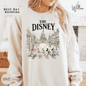 A white sweatshirt with a graphic design featuring Disney characters crossing an iconic street in front of a castle.