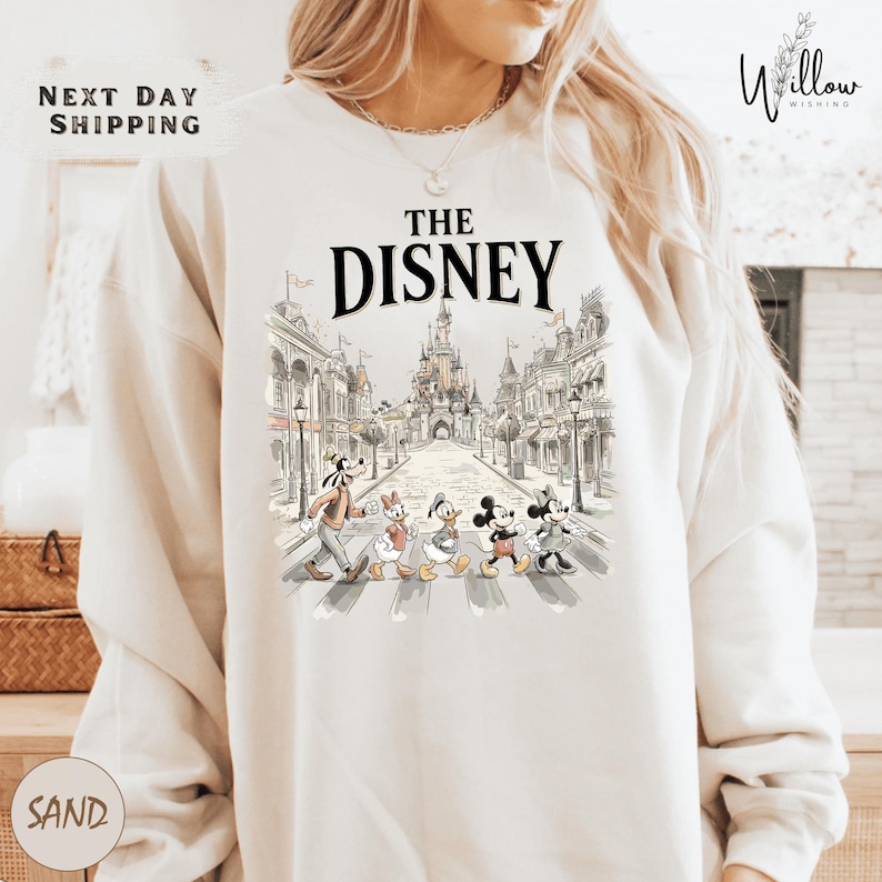 A white sweatshirt with a graphic design featuring Disney characters crossing an iconic street in front of a castle.