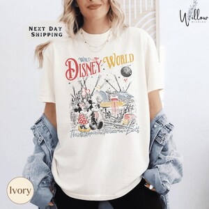 A woman wearing a white t-shirt with a Disney World graphic and text, along with a denim jacket, standing in front of a white wall.