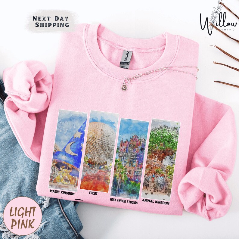 A pink sweatshirt with four colorful images of Disney landmarks printed on it, including the Eiffel Tower, Walt Disney World, Hollywood Studios, and Animal Kingdom.