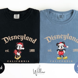 two t-shirts, one black and one blue, both featuring the iconic Mickey Mouse character from Disneyland. The black t-shirt has the text "Disneyland est. 1955" and the blue t-shirt has the text "Disneyland est. 1955".