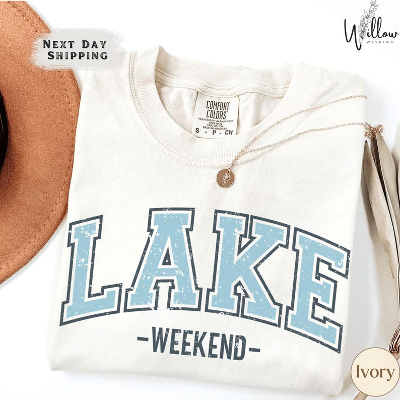 A white t-shirt with the word "LAKES" printed in blue letters, along with the text "WEEKEND" below it. The t-shirt is displayed on a white background, and there is a brown hat and a necklace visible in the image.