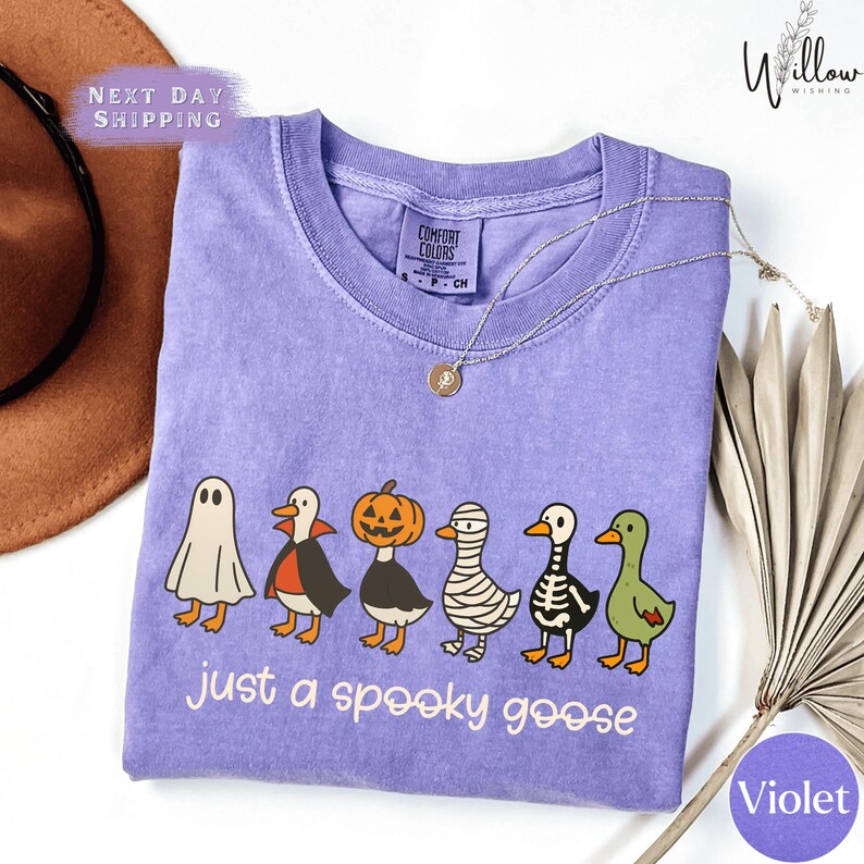 a purple t-shirt with a Halloween-themed graphic of a group of cartoon birds, including a skeleton, a pumpkin, and a ghost, along with the text "Just a spooky goose".