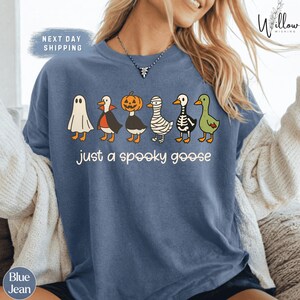 A woman wearing a blue t-shirt with a graphic design of various Halloween-themed ducks, including a ghost, pumpkin, skeleton, and green goose.