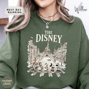 A woman wearing a green sweatshirt with a graphic of Disney characters crossing the street.