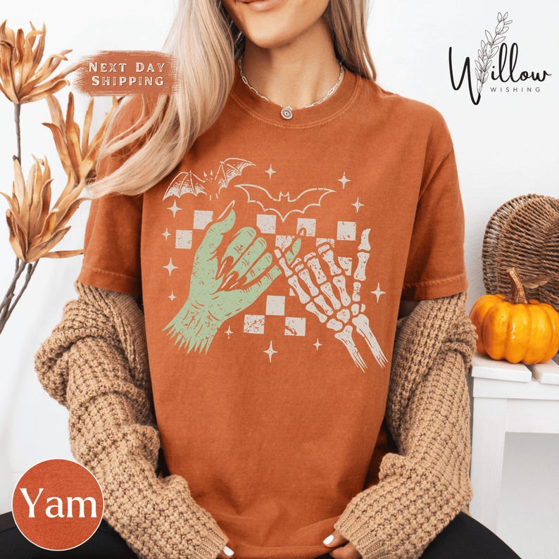 A woman wearing a brown t-shirt with a graphic design of a skeleton hand and bat on it, along with a pumpkin and a willow tree in the background.