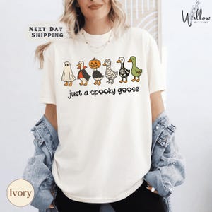 A woman wearing a white t-shirt with a graphic design of various Halloween-themed characters, including a ghost, skeleton, jack-o-lantern, and a goose, along with the text "Just a Spooky Goose".