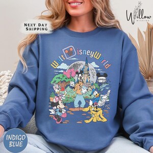 A woman wearing a blue sweatshirt with a colorful graphic of Disney characters and landmarks, including Mickey Mouse, Goofy, Donald Duck, and Cinderella Castle.