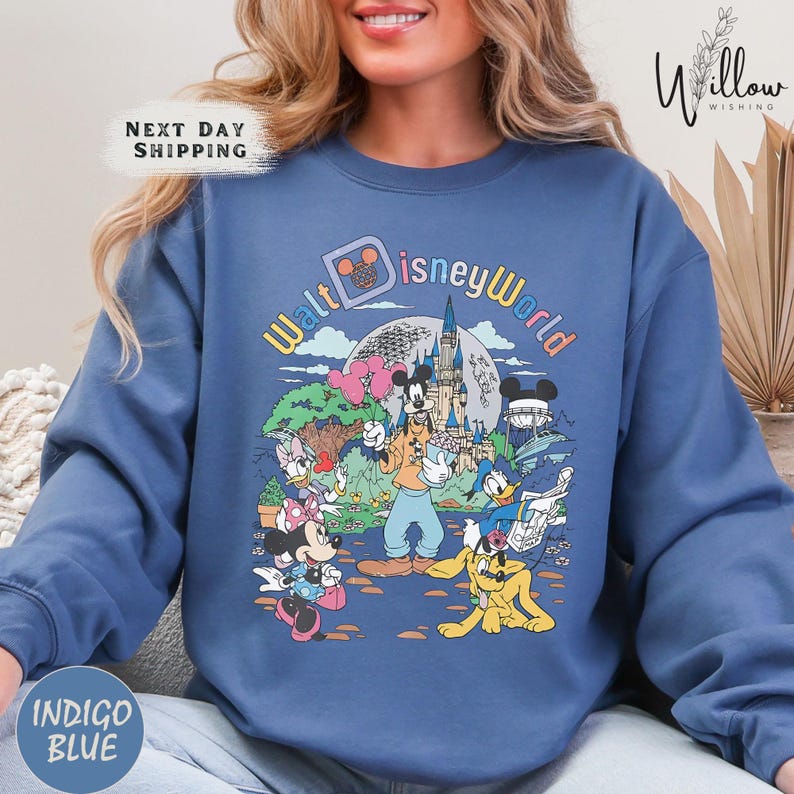 A woman wearing a blue sweatshirt with a colorful graphic of Disney characters and landmarks, including Mickey Mouse, Goofy, Donald Duck, and Cinderella Castle.