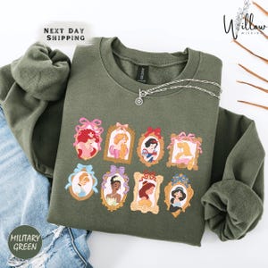 a green sweatshirt with a graphic design of various Disney princesses, including Snow White, Rapunzel, and other popular characters.