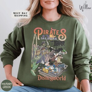 A woman wearing a green sweatshirt with a printed design of Disney characters and text related to the Pirates of the Caribbean ride at Disneyworld.
