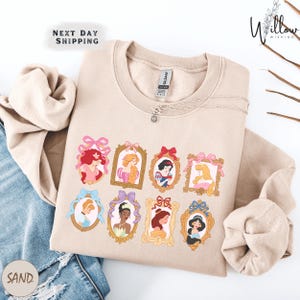 A beige sweatshirt with a graphic design featuring illustrations of various Disney princesses, including Snow White, Rapunzel, and other popular characters.