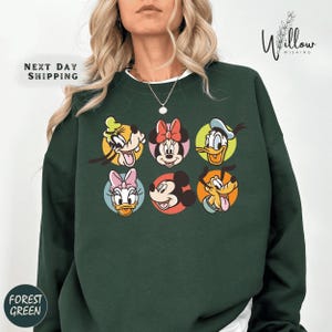 A woman wearing a green sweatshirt with a graphic of six Disney character faces, including Minnie Mouse, Donald Duck, Goofy, Mickey Mouse, Minnie Mouse, and Donald Duck.