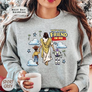 A woman wearing a gray sweatshirt with a graphic design depicting a cartoon-style illustration of a person holding a toy robot and a child, along with the text "You've got a friend in me".