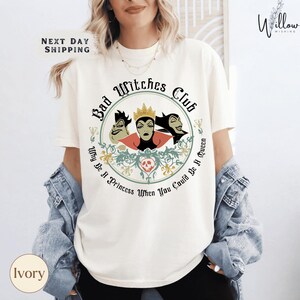 A woman wearing a white t-shirt with a graphic design featuring three cartoonish female characters and text.