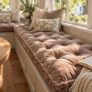 May include: A long, tufted, taupe velvet bench cushion with a thick, piped edge. The cushion is adorned with several throw pillows, including two solid taupe square pillows and a rectangular cream pillow featuring a small green leaf pattern.