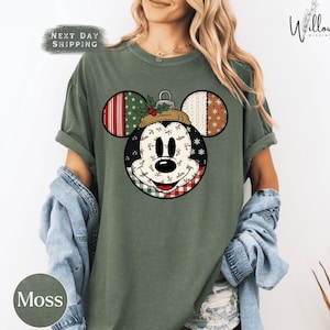 May include: Moss green t-shirt featuring a Mickey Mouse ornament design. The ornament has a patchwork design with red, green, white, and brown patterns. The text "NEXT DAY SHIPPING" is visible on the top left.