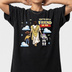 A black t-shirt with a graphic design featuring a person in a yellow robe, a cowboy hat, and a robot, along with the text "You've got a FRIEND in me" and various other cartoon-style elements.