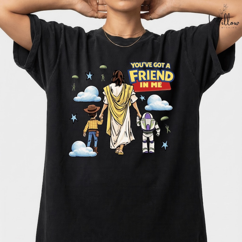 A black t-shirt with a graphic design featuring a person in a yellow robe, a cowboy hat, and a robot, along with the text "You've got a FRIEND in me" and various other cartoon-style elements.