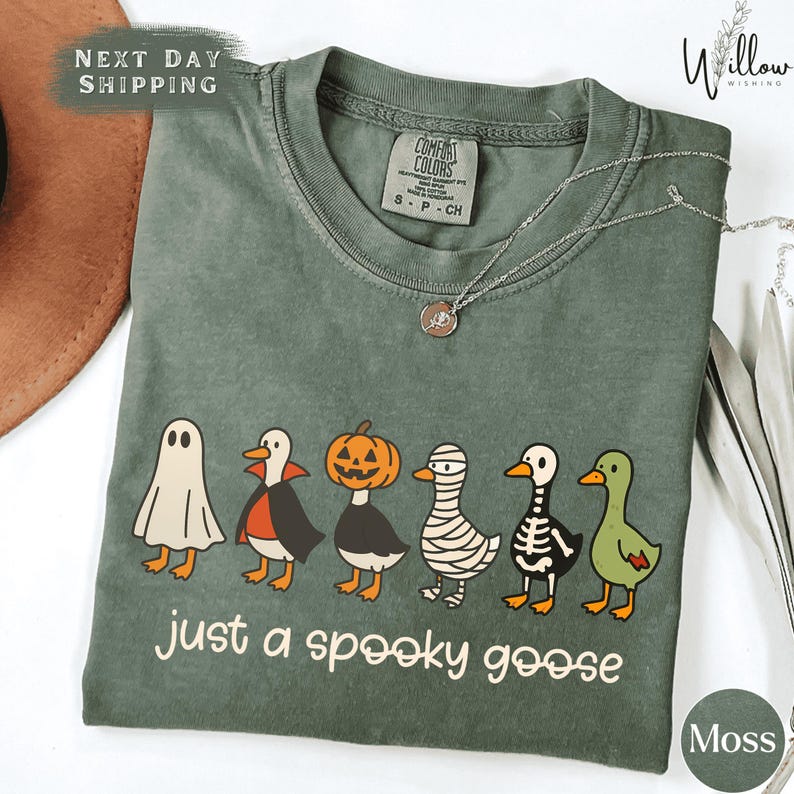a green t-shirt with a Halloween-themed graphic design, including a skeleton, a jack-o-lantern, and a ghost. The text on the t-shirt says "Just a spooky goose".