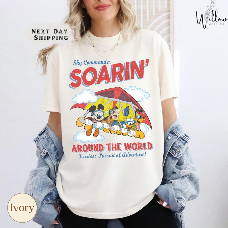 A woman wearing a white t-shirt with a graphic design of Mickey Mouse and Donald Duck on a red and yellow airplane, along with the text "Sky Commander SOARIN' AROUND THE WORLD Fearless Pursuit of Adventure!"