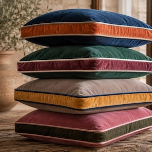May include: A stack of four square velvet throw pillows. The top pillow is navy blue with an orange side panel. The second is dark green with a maroon side panel. The third is taupe with a mustard yellow side panel. The bottom is mauve with an olive green side panel.